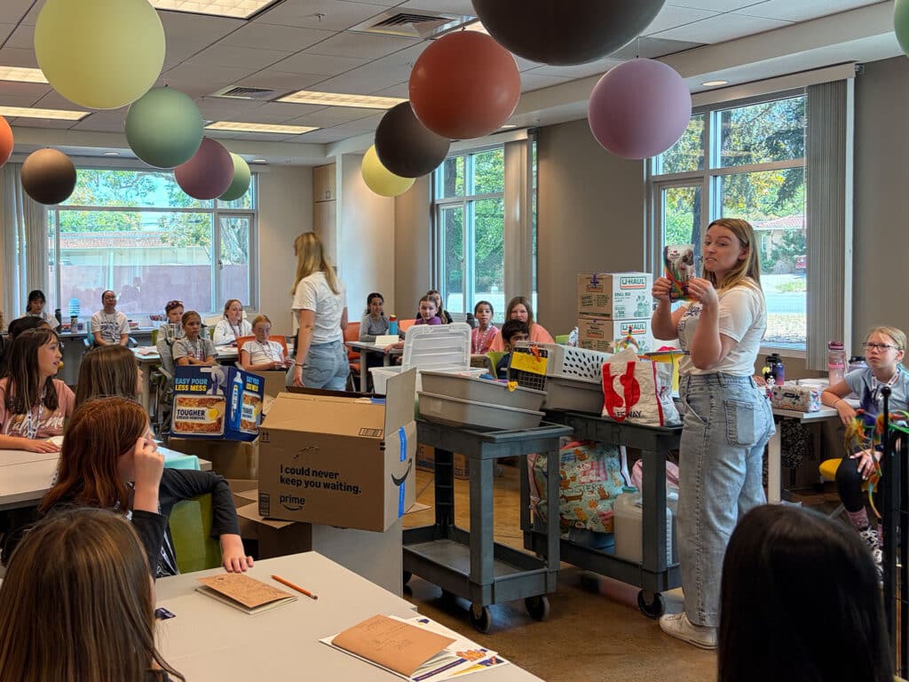 A group of young girls sits at tables in a brightly lit room equipped with large windows. One girl stands in front, speaking while another girl beside her gestures. Various boxes and supplies are organized on a cart nearby. Colorful balloons are hung from the ceiling.