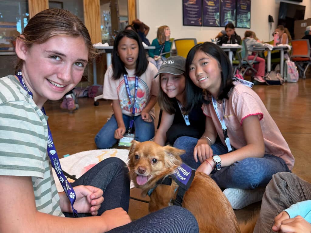 Five children are sitting on the floor in a room, smiling at the camera. They are surrounded by other children and furniture in the background. A small brown dog wearing a harness is sitting in front of them, also looking at the camera.