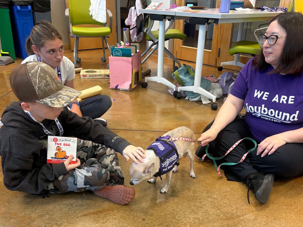 A boy wearing a camouflage cap and holding a book pets a small dog wearing a service vest, while a woman in a purple shirt kneels beside him. A girl sitting behind them looks on, and a colorful classroom with tables and chairs is visible in the background.