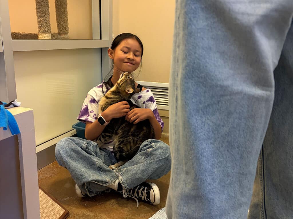 A girl sitting on the floor in casual clothing, holding a cat close to her chest. The room features a cat tree and light-colored walls, with part of a person’s legs visible in the foreground.
