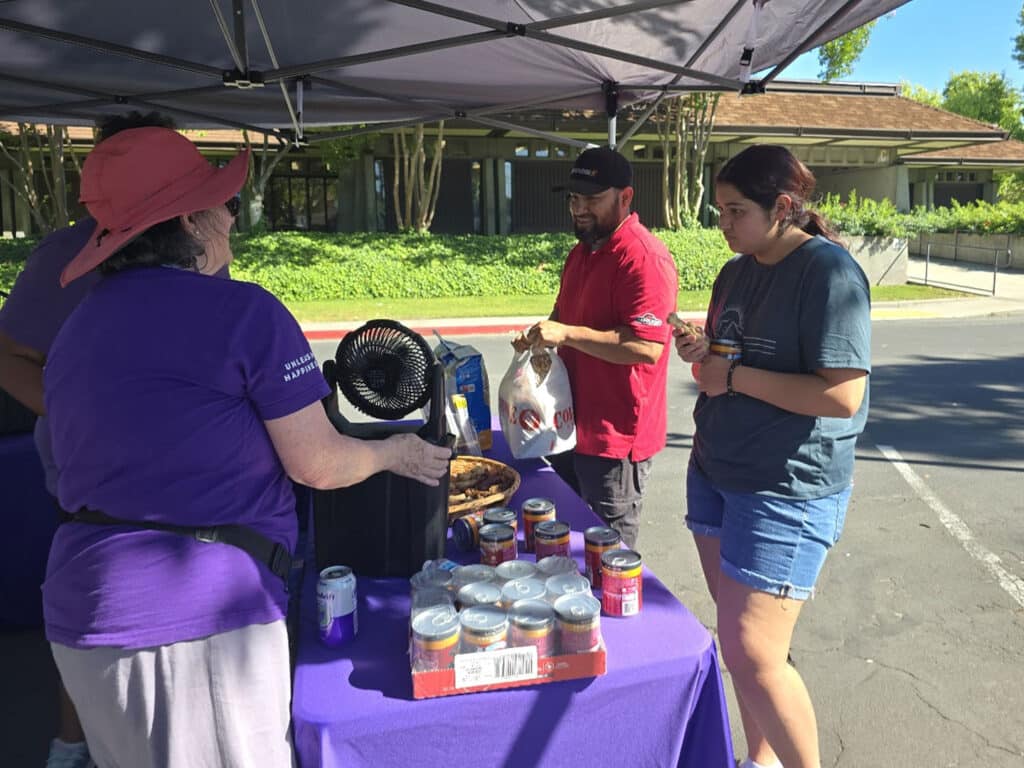 A woman in a purple shirt and hat stands behind a table, speaking to a man in a red shirt. A young woman in a gray shirt stands on the right, holding a bag. The table displays cans and a fan, and a building with greenery is visible in the background.