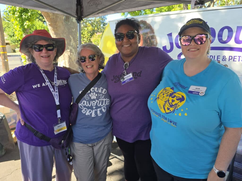 Four women stand together under a tent at a community event. They are wearing casual t-shirts and sunglasses, with one wearing a hat. A banner in the background displays the words "Joyful Rescue" and features drawings of pets.