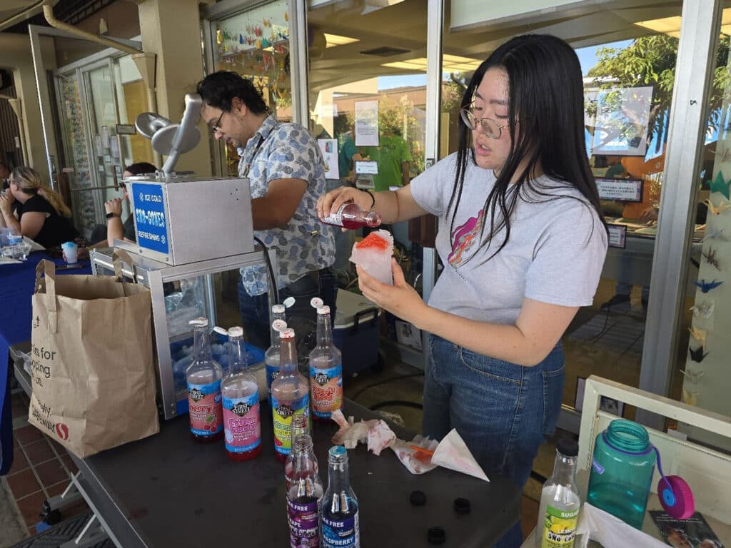 A young woman with long hair in glasses pours syrup onto a shaved ice treat at a food stand. Colorful bottles of soda and a brown paper bag are placed on the table, with a man in a patterned shirt in the background, engaged in conversation.