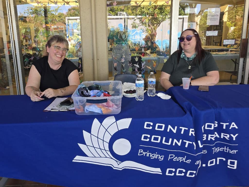 Two women sit at a table covered with a blue cloth displaying the Contra Costa County Library logo. The table has a container of items, two water bottles, and a pink drink. In the background, there are large windows and colorful decorations.