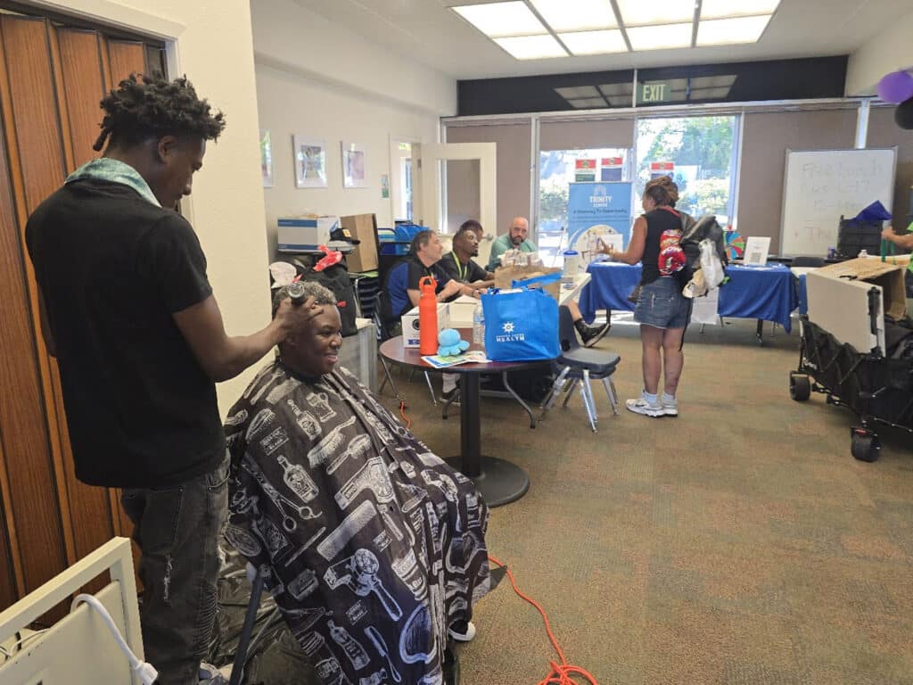 A young man with a dark shirt cuts the hair of a seated man wearing a patterned cape in a community room. Others are engaged in various activities at tables in the background, surrounded by chairs and supplies.