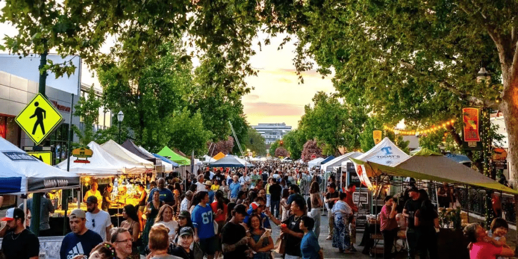 A bustling street scene at Oakley Summerfest with numerous food and craft vendor tents lining both sides. A large crowd mingles along the tree-lined street, with greenery and rustic lights adding to the festive atmosphere.