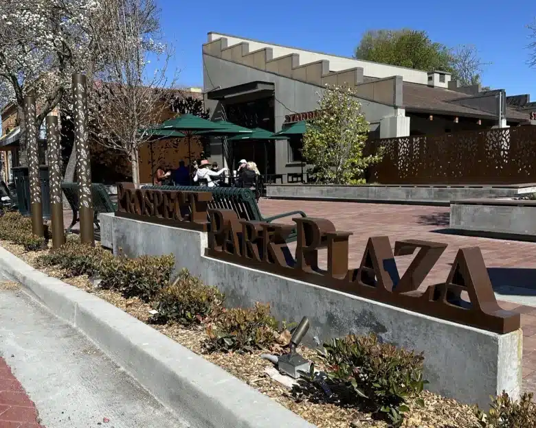 A sign for 'Museum Park Plaza' made of metal is mounted on a concrete base, surrounded by landscaped shrubs and flowers. In the background, outdoor tables and chairs are visible under green umbrellas, with a building featuring a modern design.