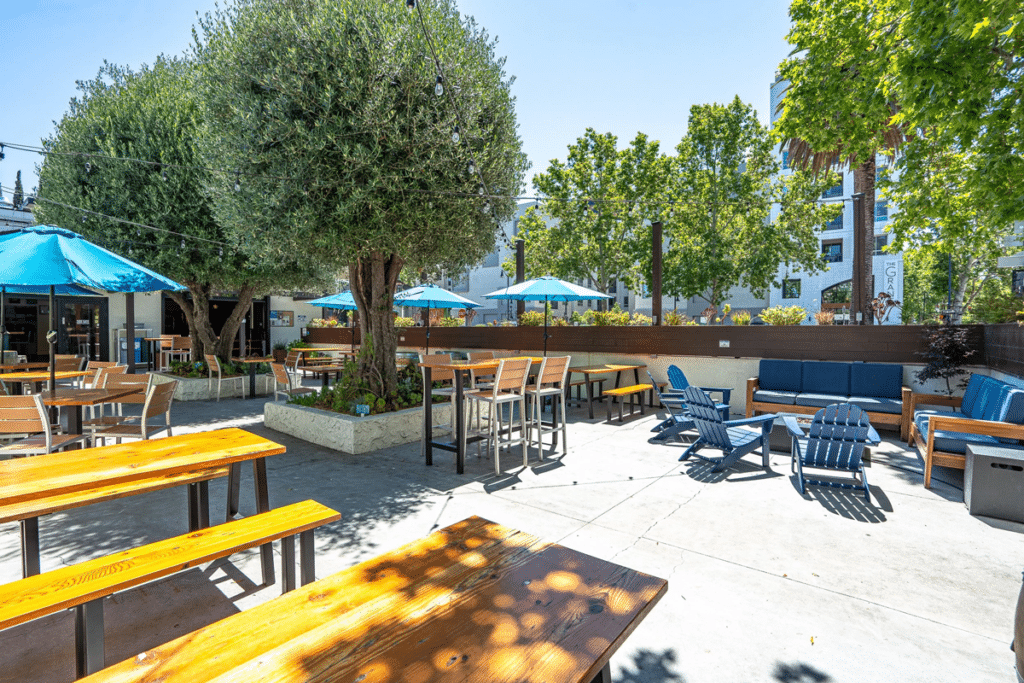 An outdoor seating area featuring wooden tables and chairs under blue umbrellas. There are several trees providing shade, and a cozy seating arrangement with blue lounge chairs is visible against a backdrop of a modern building.