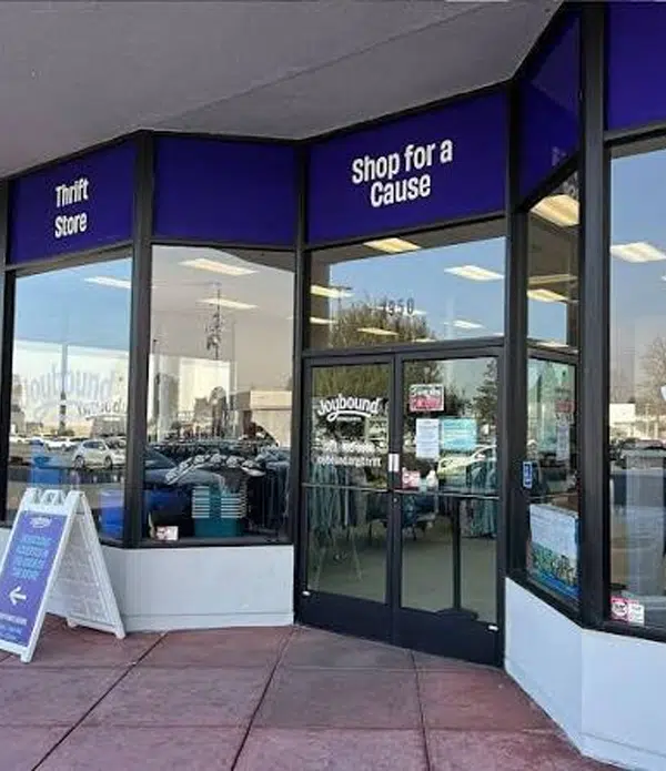 The entrance of a thrift store features large windows and a black door. Above the door, a sign reads "Shop for a Cause" on a purple background. A white sign nearby advertises the store's hours, alongside parking visible in the background.