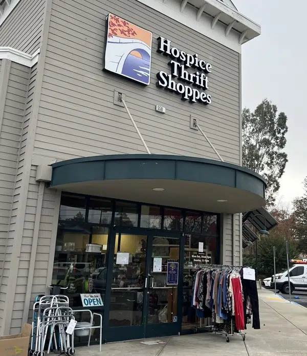 Exterior view of the Hospice Thrift Shoppes storefront featuring a blue awning and a large sign. The entrance has glass doors, with clothing racks displaying various garments to the right and a parking lot visible in the background.