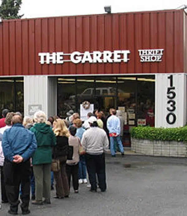 A crowd of people is waiting in line outside a thrift shop named "The Garret," located at 1530. The shop has large windows displaying items inside, and the exterior is painted in red and white.