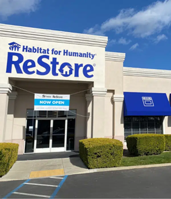 The exterior of a Habitat for Humanity ReStore building, featuring a large sign that reads "ReStore" and smaller text that says "Habitat for Humanity." The entrance has glass doors with a banner stating "NOW OPEN". A blue awning is visible on one window.