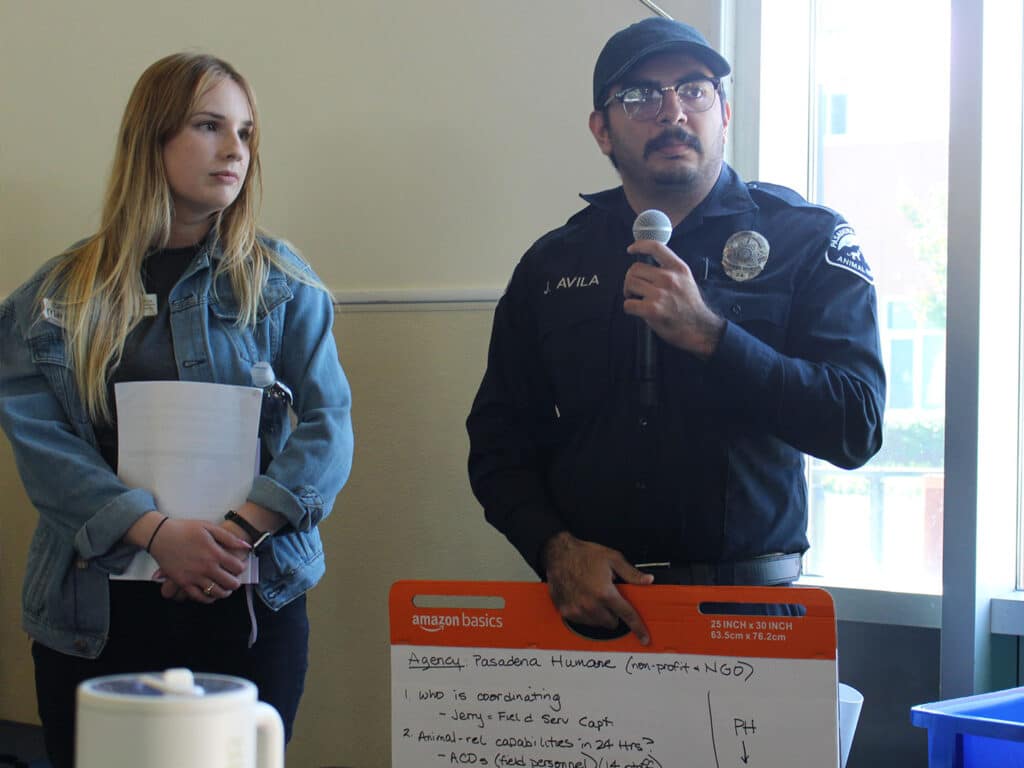 A man in a police uniform speaking into a microphone stands beside a woman with long blonde hair wearing a denim jacket. A whiteboard displaying written notes is positioned in front of them, with a kettle and blue containers visible in the foreground.