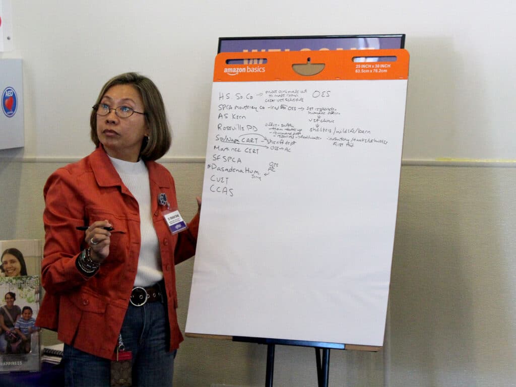 A woman with shoulder-length brown hair wearing glasses and an orange jacket stands next to a whiteboard filled with handwritten notes and bullet points. The background features a bulletin board with various materials.