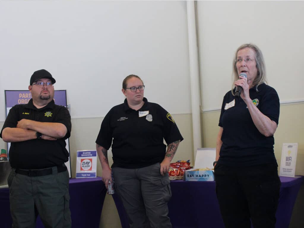 Three emergency response professionals are standing in front of a display table at a workshop. A man in a black cap and a sheriff's badge is on the left, a woman in a black uniform is in the center, and a woman speaking into a microphone is on the right, wearing a dark shirt with a logo.
