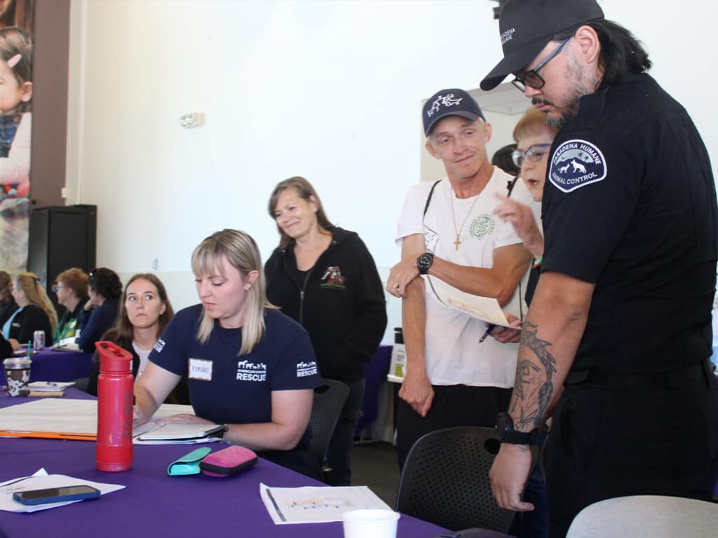 Participants engaged in a discussion at a disaster response workshop. A woman in a blue shirt takes notes at a table, while another woman stands beside a man in uniform, who appears to be providing guidance. The setting includes purple tablecloths and various materials on the tables.