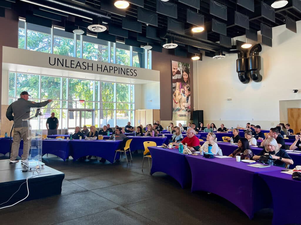 A speaker gestures towards the audience in a brightly lit room with large windows. Attendees are seated at tables covered with purple cloths, listening attentively during the Disaster Response Workshop, with the phrase "UNLEASH HAPPINESS" displayed above.