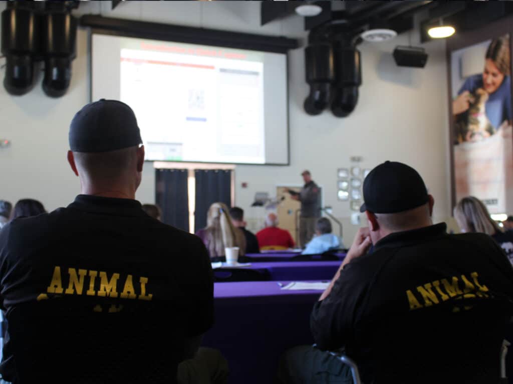 Two individuals in black shirts with the word "ANIMAL" displayed in yellow sit in a classroom setting, facing a presenter at the front. A screen displays information, while other attendees are partially visible in the background.