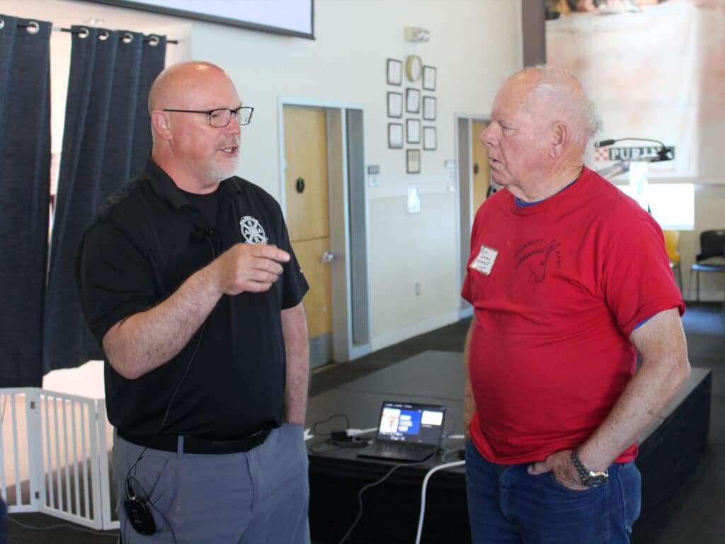 A man in a black polo shirt and glasses talks to an older man wearing a red shirt. The background includes a laptop on a table and curtains, with some framed pictures visible on the wall.