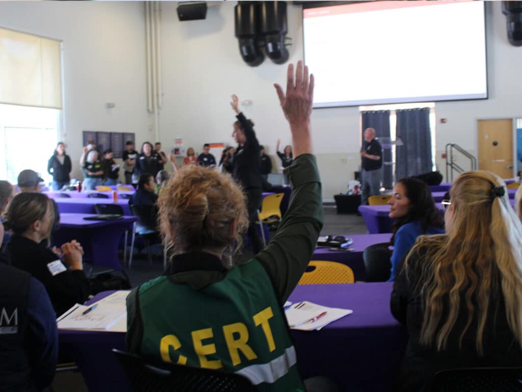 A group of participants at a disaster response workshop is seated at tables covered with purple tablecloths. An attendee wearing a green "CERT" vest raises her hand while facing a speaker in front of a presentation screen.