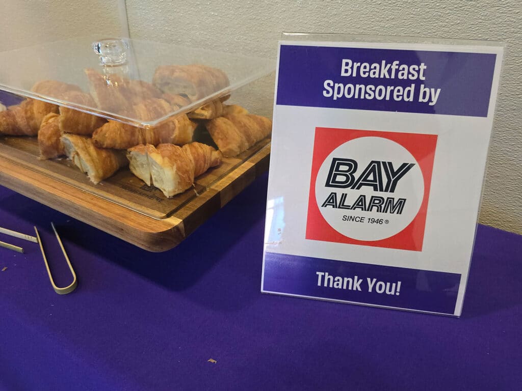 A display featuring a clear container of croissants on a wooden platter beside a sign that reads, 'Breakfast Sponsored by BAY ALARM Since 1946. Thank You!' The backdrop is a purple tablecloth.