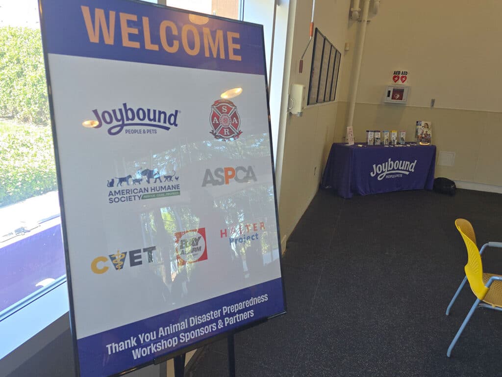 A welcome sign featuring logos from several organizations, including Joybound, American Humane Society, ASPCA, CVET, and Fear Free, displayed in a room with natural light. A table with brochures and materials is visible in the background.