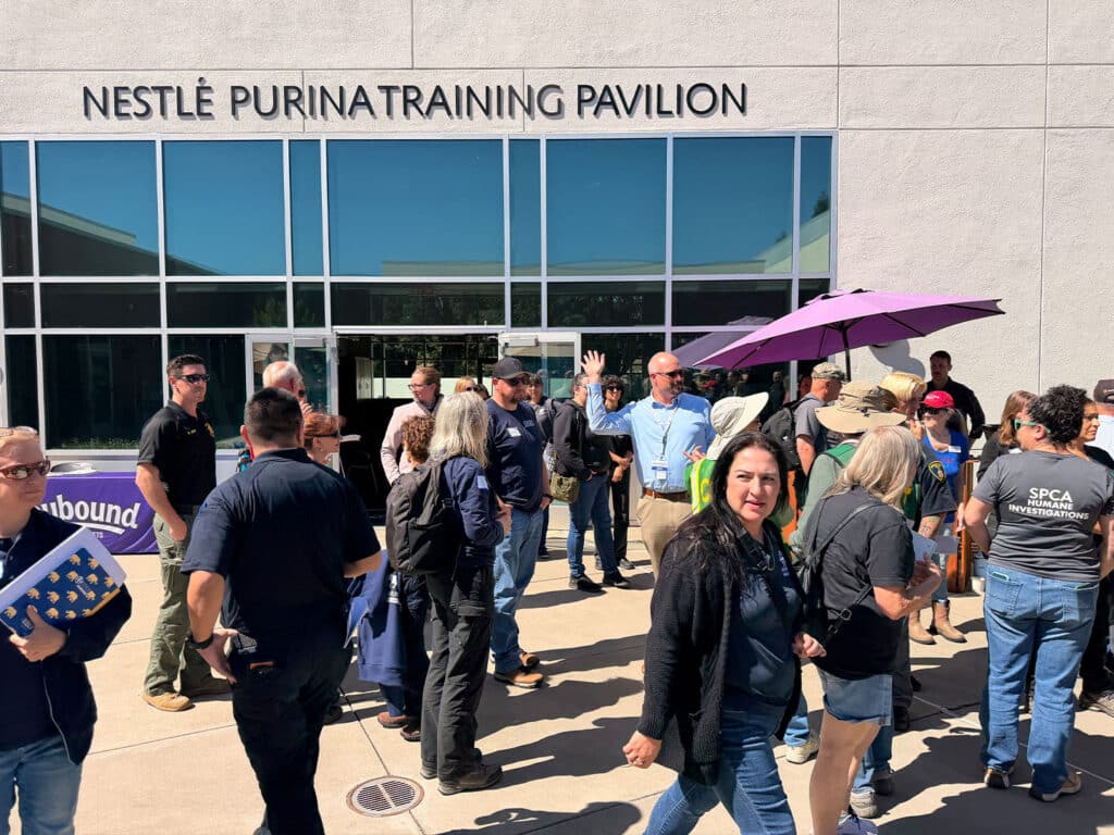 A diverse group of people gathered outside the Nestlé Purina Training Pavilion. Some individuals hold umbrellas, while others engage in conversation, with a clear view of the building's large windows and signage above the entrance.