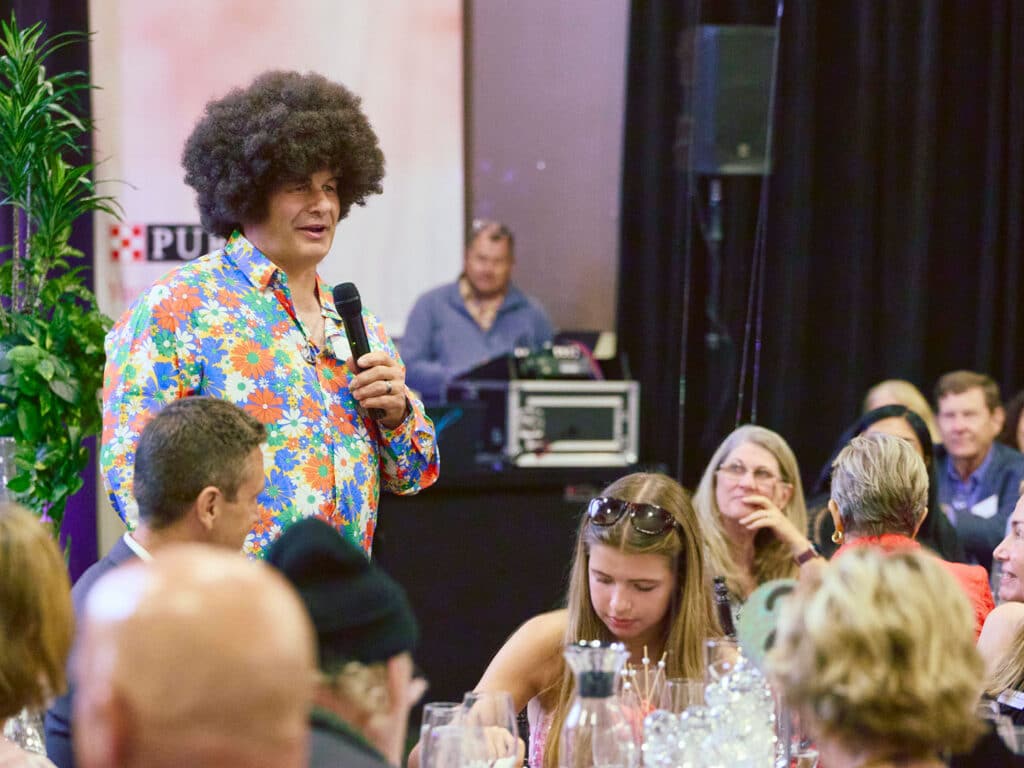 A man with curly hair, wearing a colorful floral shirt, speaks into a microphone while standing in front of seated guests at a gala event. Several attendees, including a woman with long hair and a girl, are listening attentively.