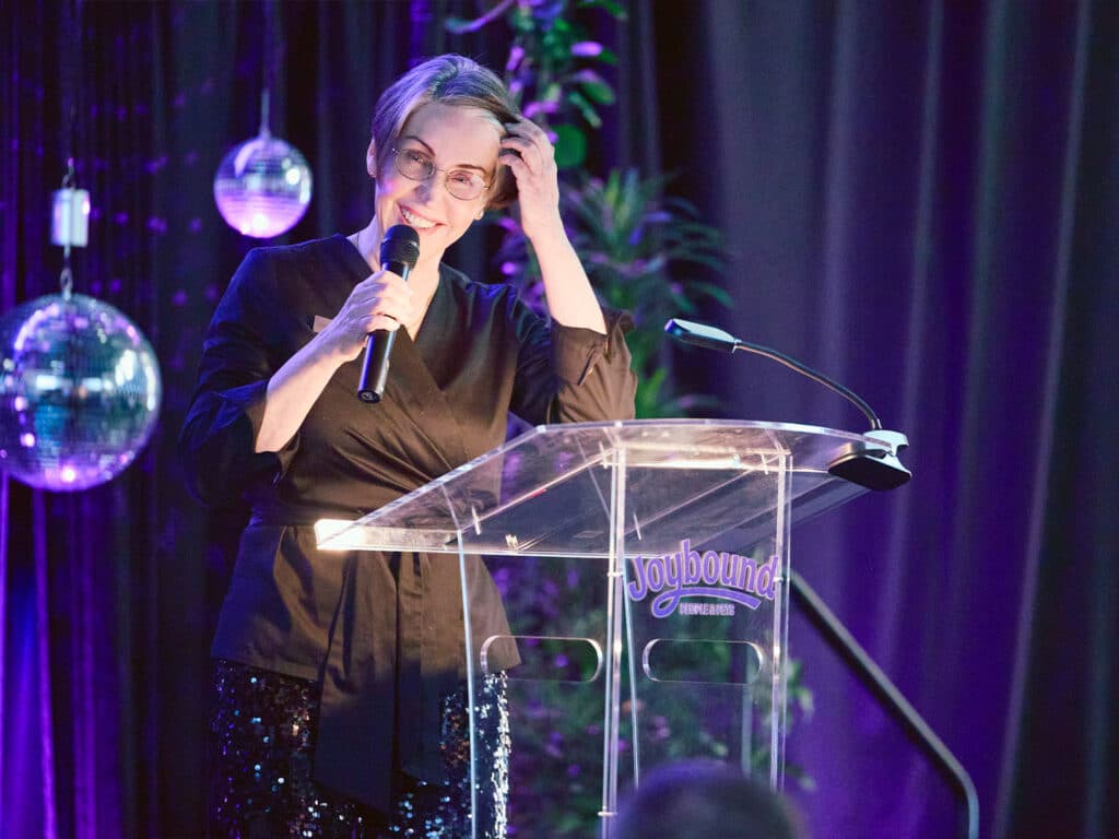 A woman with short gray hair and glasses stands at a clear podium, holding a microphone. She is wearing a dark blouse and is speaking in front of a black backdrop adorned with disco balls and greenery.