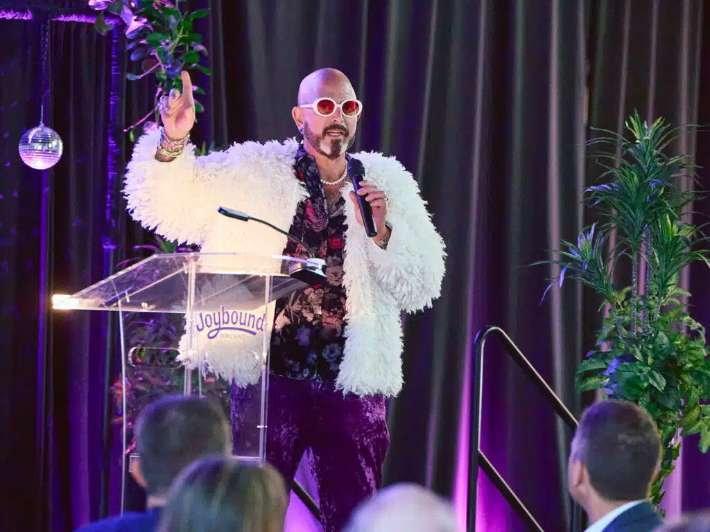 A man wearing large sunglasses and a white faux fur coat speaks into a microphone from behind a clear podium with the word 'Joybound.' He is gesturing with one hand, while the audience sits in front of him, with potted plants visible in the background.