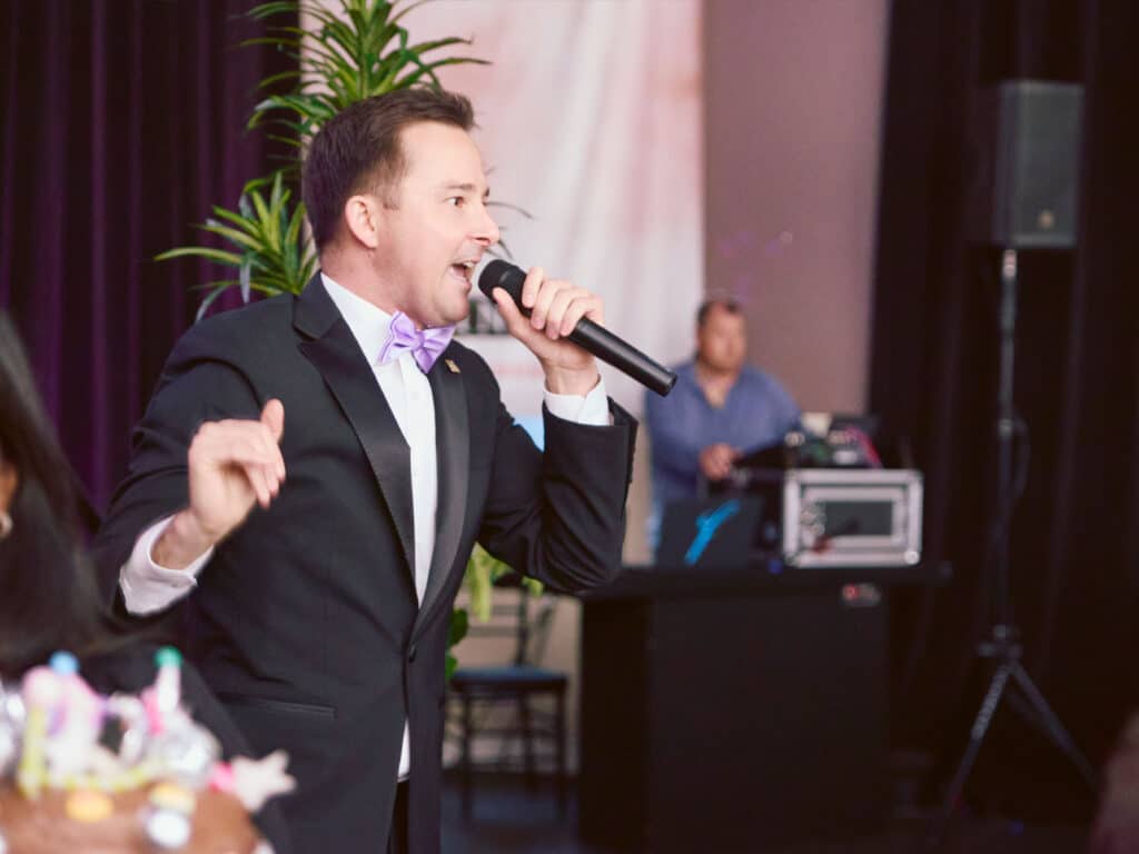 A man in a black tuxedo and purple bow tie speaks into a microphone while gesturing. He is in front of a decorative background featuring plants, with a DJ setup visible in the background.