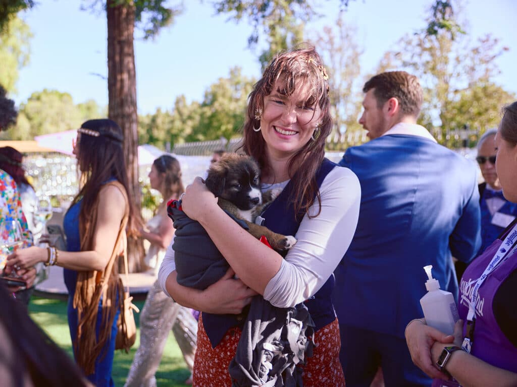 A woman with long brown hair carries a small brown and white puppy in her arms at an outdoor event with people in the background. The scene includes trees and greenery, suggesting a sunny day.