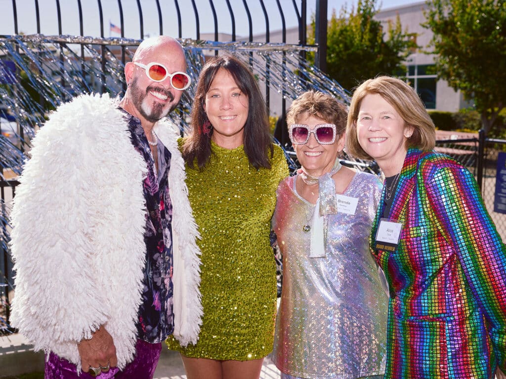 Four individuals pose together outdoors, wearing vibrant, sequined outfits. The man on the left wears a white fluffy jacket and glasses, while the woman in the center dons a shiny green dress. The other two women are dressed in reflective dresses with colorful patterns. A fence and trees are visible in the background.