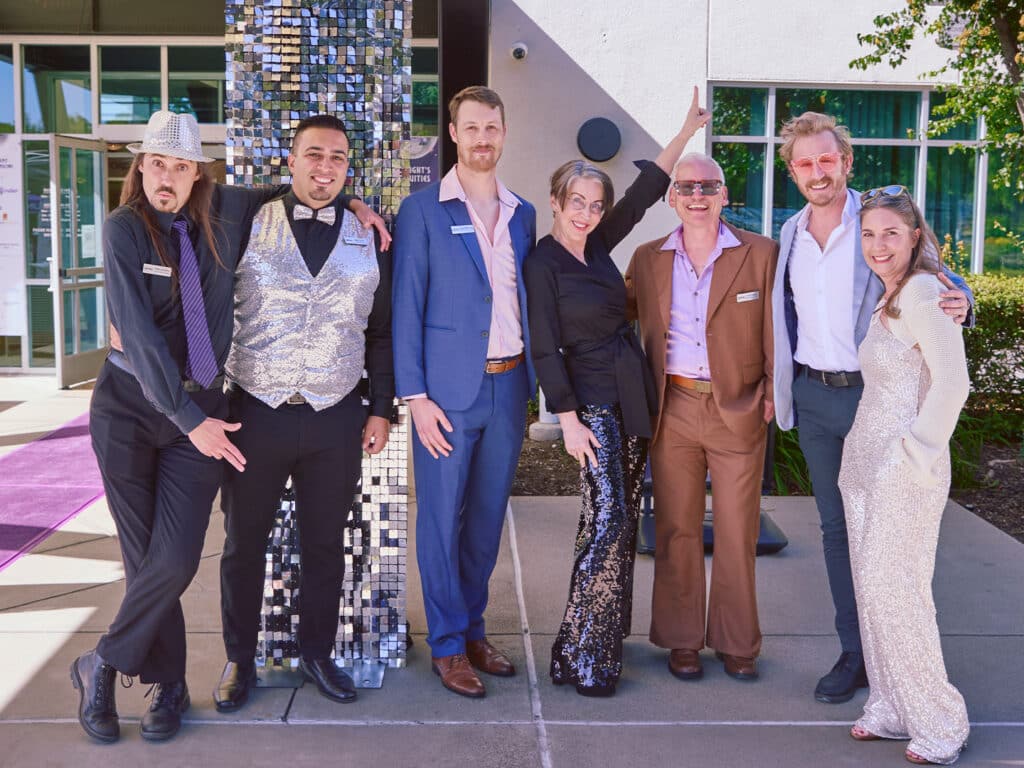 Seven individuals pose for a photo outside a building adorned with a reflective mosaic column. They wear formal attire, including suits and dresses, and stand against a backdrop featuring large windows and greenery.