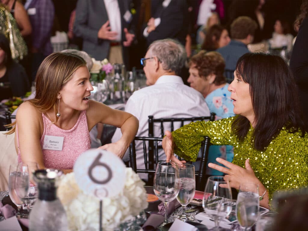 Two women engage in conversation at a dining table during the Unleash Happiness Gala. The woman on the left wears a pink dress and the woman on the right is in a green sequined outfit; both are surrounded by dinnerware and floral centerpieces.