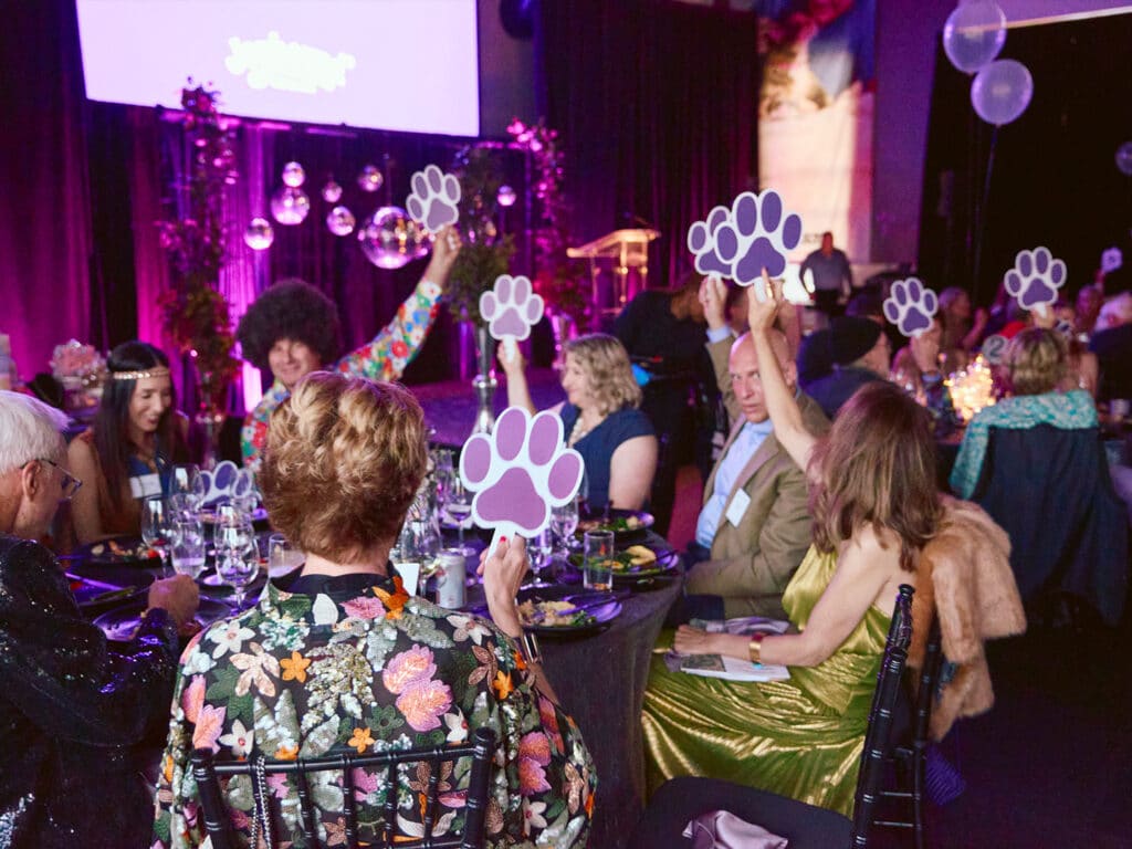 A group of people seated at round tables during an event, each holding up signs shaped like paw prints. The background features purple lighting and decorations, with a stage area and large screens visible.