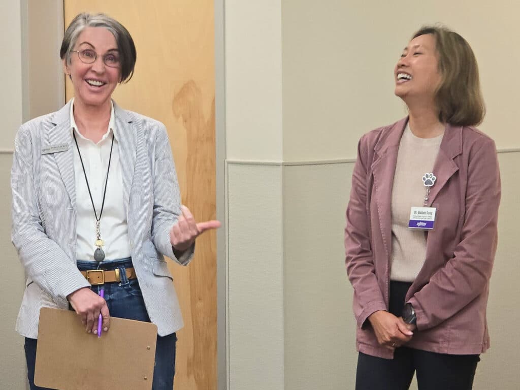 Two women stand facing each other in a room. One woman, wearing a gray blazer and holding a clipboard, gestures with her right hand while speaking. The other woman, wearing a light pink blazer and a nametag, appears to be laughing. The background features a beige wall.