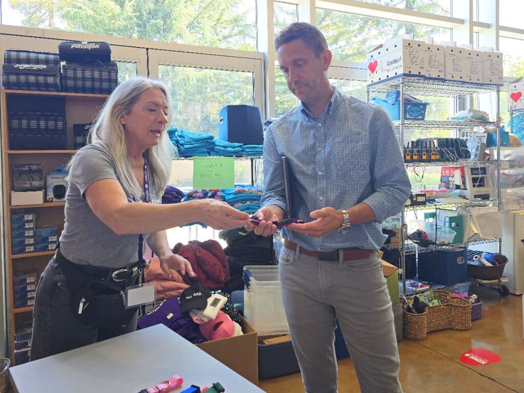 A woman with long gray hair wearing a gray shirt hands an item to a man in a blue checkered shirt at a counter. The background includes shelves with various items and bright natural light coming through large windows.