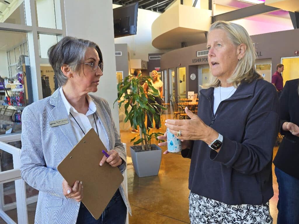 A woman with short gray hair and glasses, holding a clipboard, stands beside another woman with long blonde hair, discussing while holding a cup. The background features a bright, open space with tables and plants.