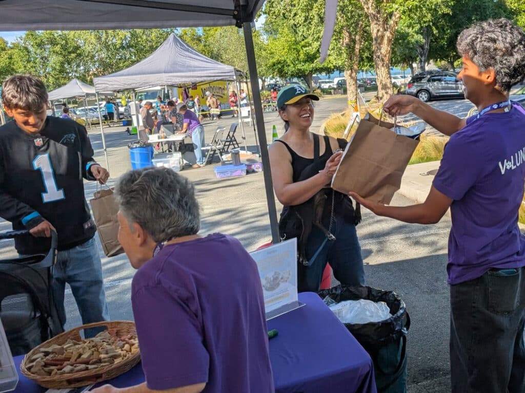 A man in a black football jersey and another man in a purple shirt stand near a table where a woman in a black cap hands a paper bag to a person with gray hair. The background shows multiple tents and people at an outdoor event.