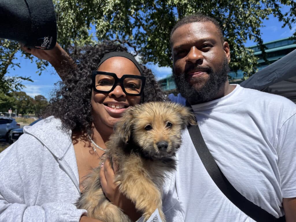 A woman wearing large black glasses and a light sweater holds a small, fluffy puppy, smiling alongside a man with a beard wearing a white t-shirt. They are standing outdoors with trees and a clear sky visible in the background.