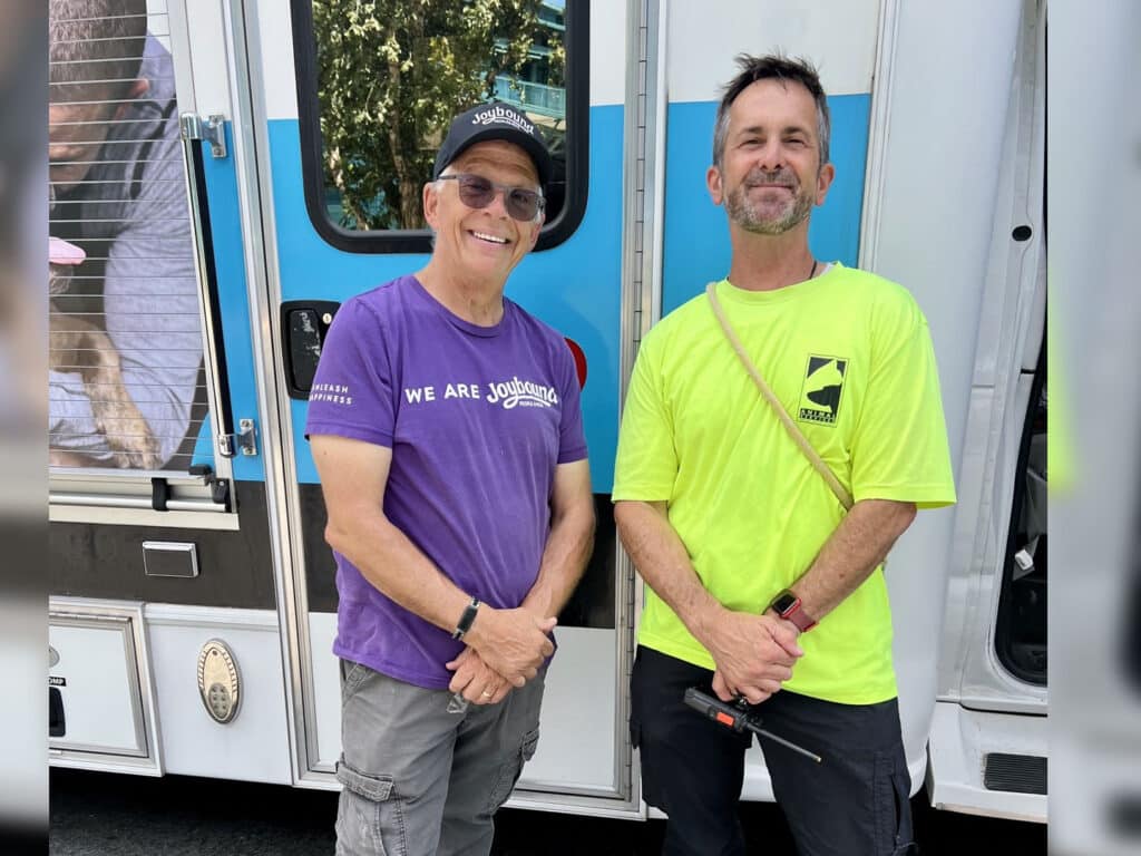 Two men stand outside a vehicle in a bright outdoor setting. One man is wearing a purple shirt that says "WE ARE JONATHAN" and gray shorts, while the other is wearing a neon yellow shirt and black pants, holding a tool.