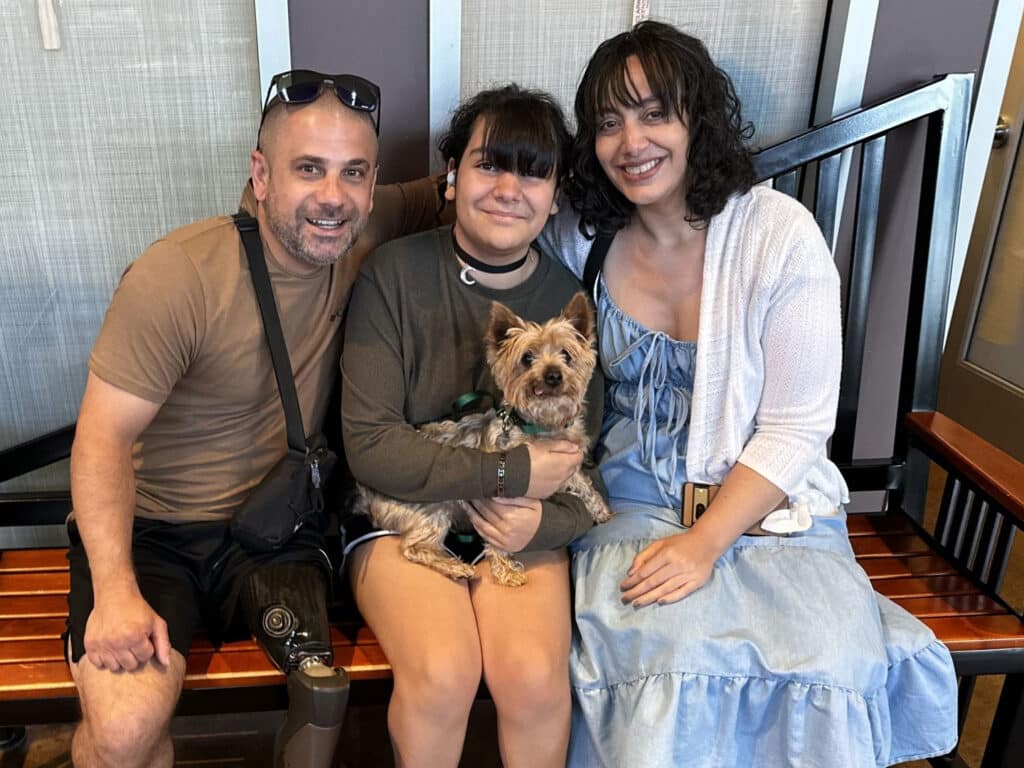 A man wearing sunglasses and a brown shirt sits on a bench beside a teenage girl holding a small dog, while a woman in a white cardigan sits on the other side. The setting features a modern interior with silver accents.