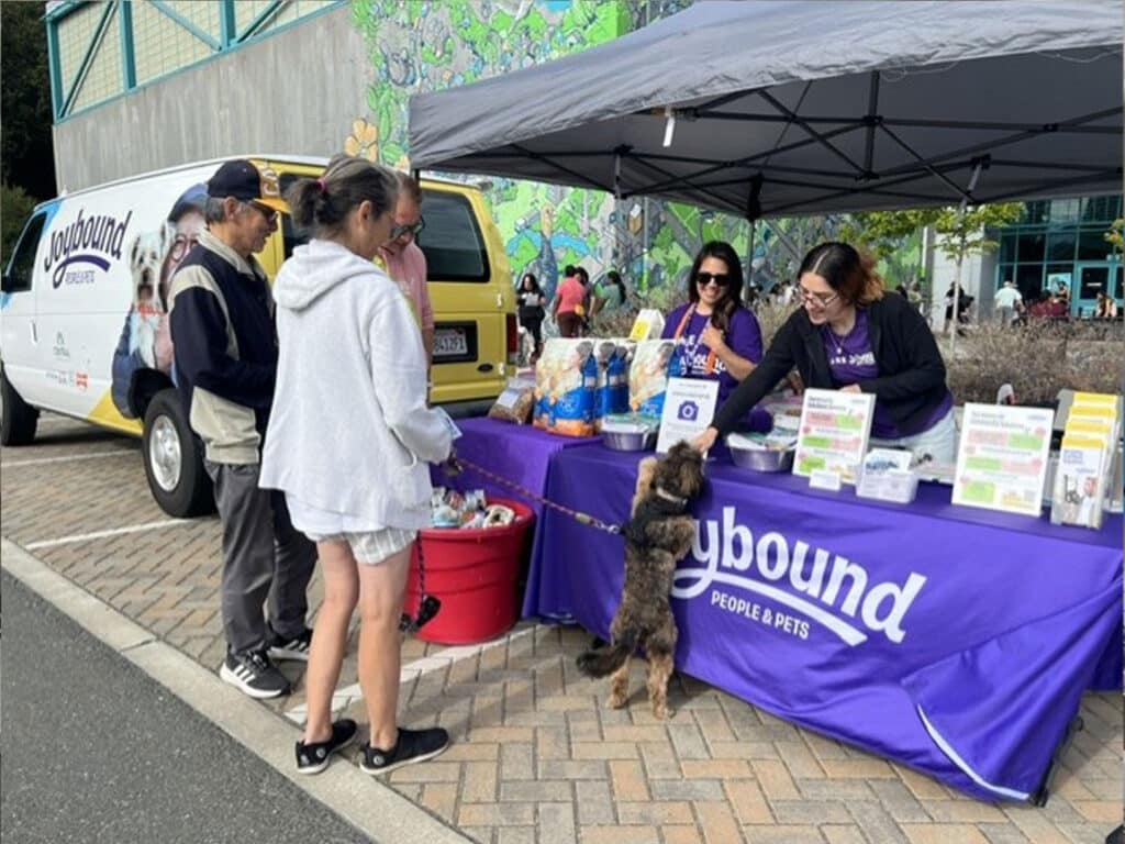 A booth promoting pet resources features a range of pet supplies on a table. Two women assist visitors while a dog interacts with a person. In the background, a van with the logo 'Playbound People & Pets' is parked beside a colorful mural.