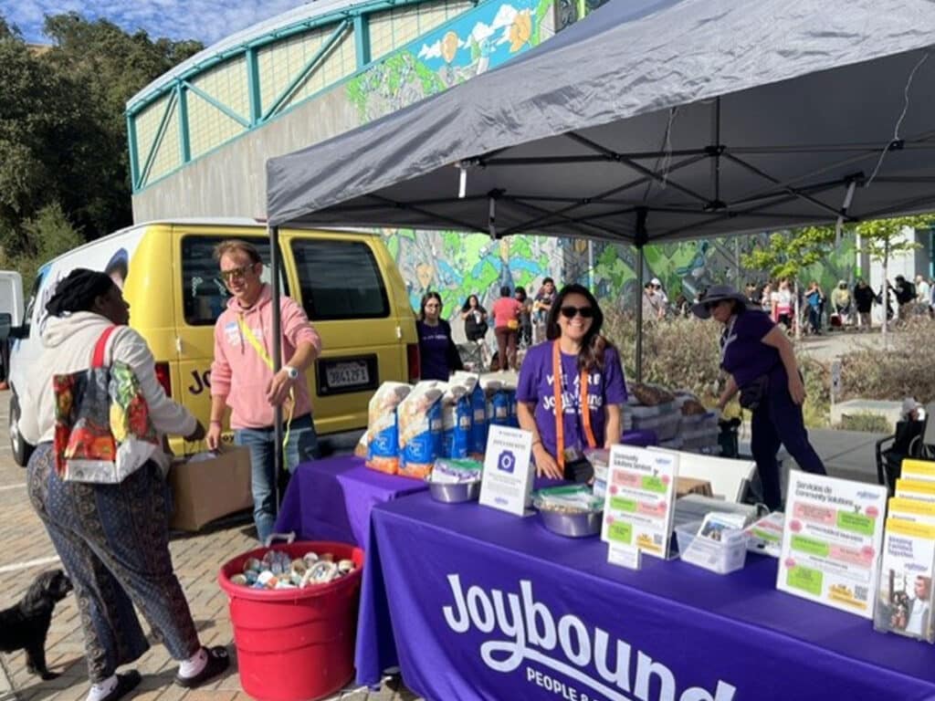 A woman wearing a purple shirt stands behind a table with food items and informational flyers at a community food pantry event. A yellow food van is parked nearby, and several people are interacting around the setup under a tent.