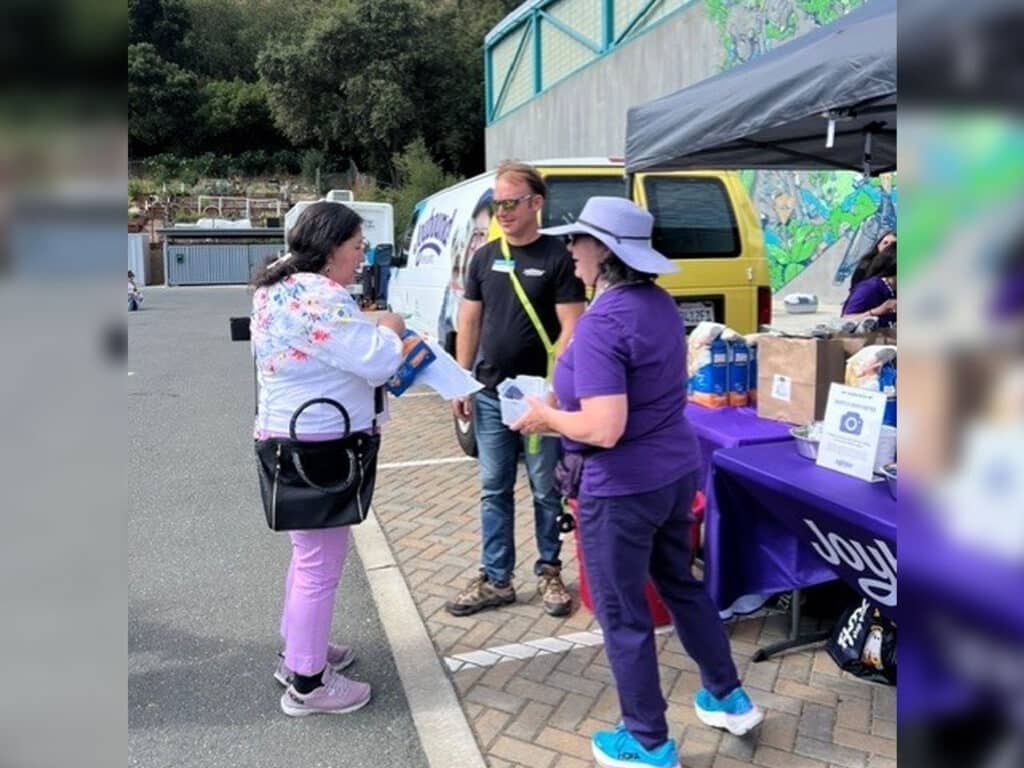 A woman in a white floral sweater interacts with a man wearing glasses and a black shirt, while another woman in a purple shirt hands out items at a FoodShare Pantry event. A yellow vehicle and a tent with supplies are in the background.