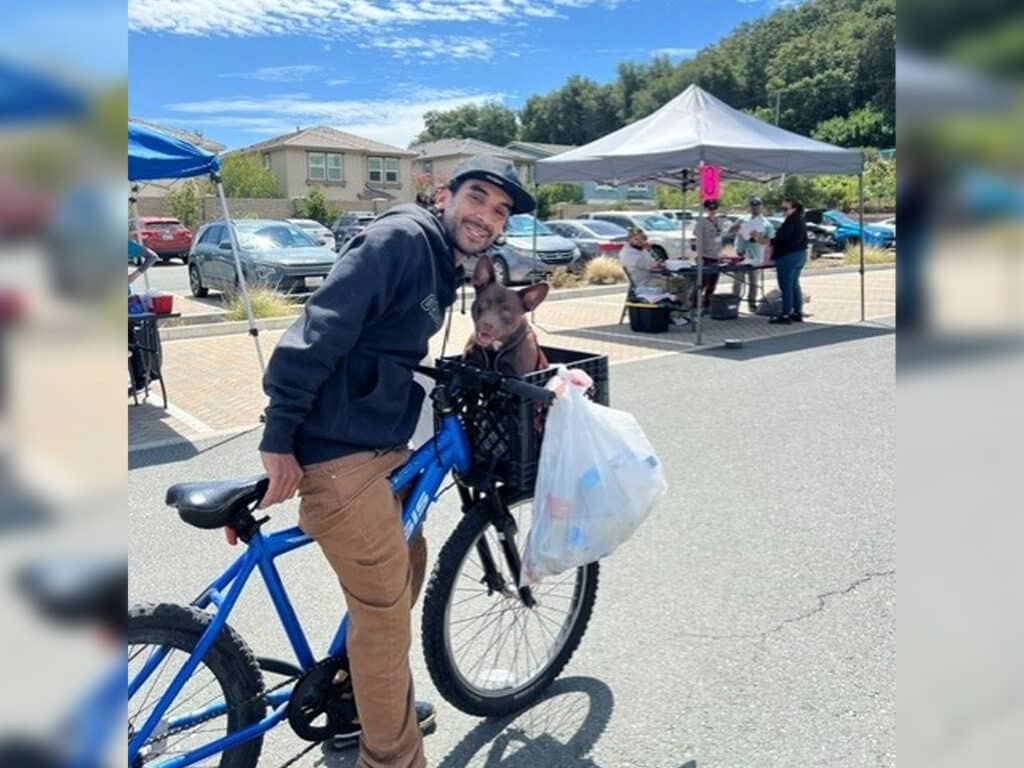 A man wearing a gray hoodie and cap is smiling while holding a plastic bag on a blue bicycle. A small brown dog sits in the bicycle's front basket. In the background, there are tents and people at an outdoor event.