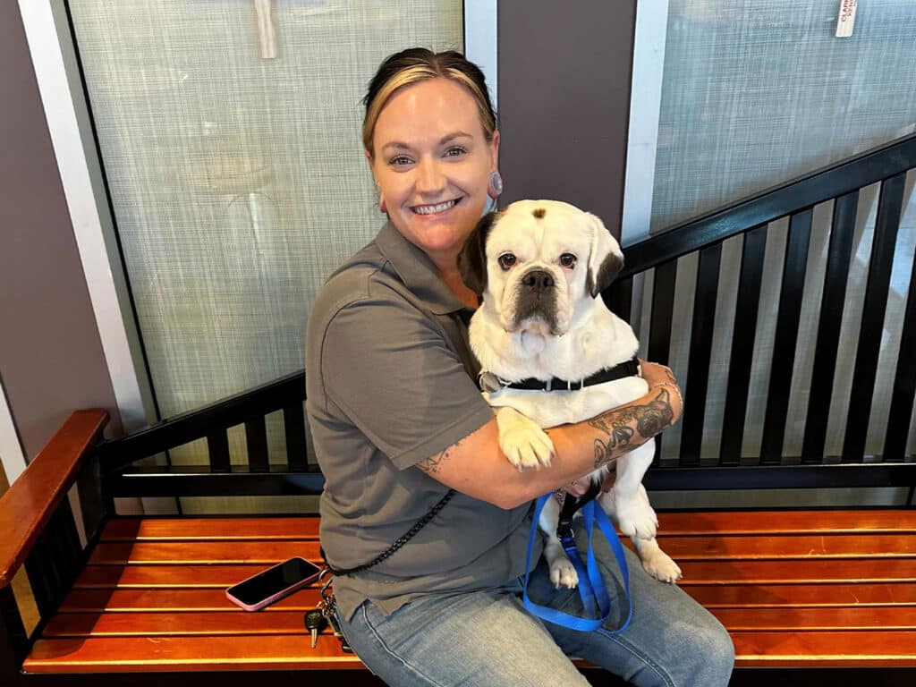 A woman with tattoos, wearing a gray shirt, holds a white dog in her lap while sitting on a wooden bench. The backdrop features large windows and a modern interior setting.