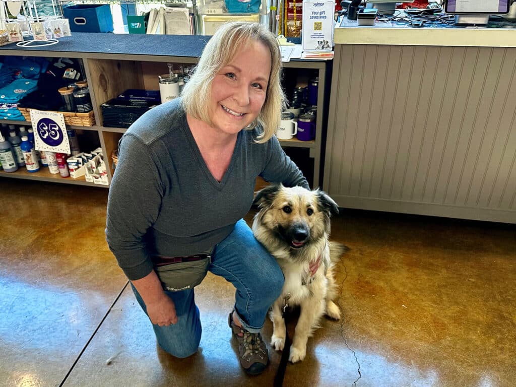A woman with blonde hair wearing a gray sweater kneels beside a dog with a tan and white coat in a pet store. The store features various items visible on shelves in the background, with warm wooden flooring.