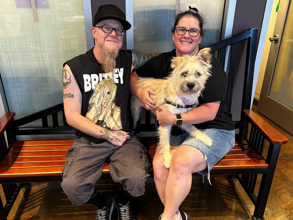 A man with glasses and a tattooed arm sits on a wooden bench beside a woman wearing glasses and a black tank top. She holds a small, light-colored terrier mix dog. The background features soft lighting and a glass wall.
